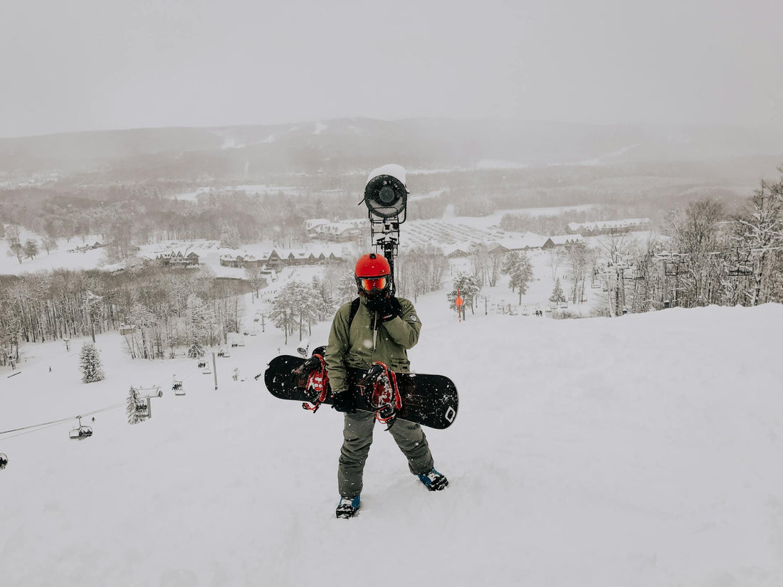 A snowboarder standing on a snowy mountain field holding their board, alpine resort visible in the background