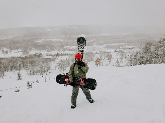 A snowboarder standing on a snowy mountain field holding their board, alpine resort visible in the background